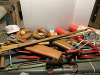 Mix of sports equipment on table including tennis rackets, baseball gloves, bats, hockey sticks, archery bows, and skate sharpener box.