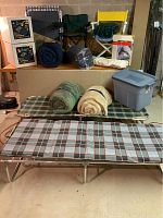 Photo showing two folded sleeping bags, two folding chairs (green and yellow), boxes with Comfortcoil air bed, rolled items, and plastic storage bin on a raised platform and floor.