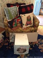 Five Christmas themed throw pillows arranged on a chair including designs of a brown bear, buffalo check pattern, a Christmas tree, and cat with stocking. Also visible is a boxed collectible house and rustic Christmas sign.