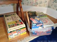 Photo showing a mixed lot of board games in original boxes including The Uncle Wiggly Game, Risk, Boggle, and Monopoly editions plus Xbox and DVD cases on a chair and plastic container.