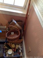 Photo shows assorted woven baskets stacked together in a corner with a wooden walking stick leaning against the wall nearby.
