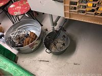 Large galvanized tub containing wire plant holder, hanging baskets, and other items on basement floor next to small floral chair.