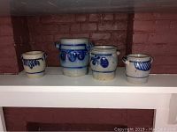 Four vintage blue and gray salt glazed pots with decorative strapping and small handles, placed on shelf against brick wall.