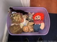 Overhead view showing multiple dolls including Raggedy Andy, Cabbage Patch, baby doll, rabbit plush and braided yarn doll head in a plastic storage bin.
