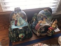 Two Disney musical snow globes side by side on a wooden table by window, one Cinderella dance scene globe with castle base, the other a multiple princess gathering globe around fountain.