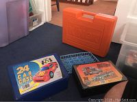 Photo of three toy storage cases on carpet: blue Matchbox 24 car case with red car graphic, orange Knex case, black Hot Wheels 24 car case with racing label. Also blue plastic car tray insert and Thomas the Tank Engine metal box visible.