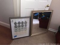 Beveled mirror beside framed wall art showing leaf design, items on floor leaning against a door.