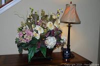 Photo showing the lamp and faux floral arrangement together on wooden surface against beige wall, lamp turned on showing lampshade light.
