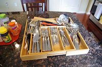 Flatware and serving utensils arranged in bamboo organizer on countertop with nearby vitamins, showing forks, spoons, knives, serving spoons, ladles, and chopsticks.