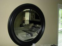 Front view of round wall mirror with thick black frame showing reflection of bedroom.