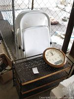 Photo showing metal shoe shelf with perforated shelves and bent top shelf, analog wall clock placed on top and 2 white folding chairs leaning behind.