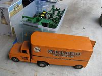 Plastic bin containing two green John Deere toy tractors with yellow wheels and various green agricultural attachments, next to a large orange Allied Van Lines toy truck.