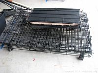 Medium sized black metal wire pet cage parts stacked flat on the floor, showing base trays and wire panels with wheels attached.