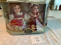 Front view of boxed porcelain twin dolls, female and male, seated and dressed in vintage-style clothing, box is visibly damaged.