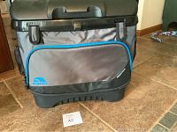 Front and side view of the large Igloo cooler showing gray fabric with blue piping, black hard plastic base, wheels, and front zipper compartment with Igloo logo.