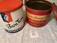 Two vintage tin cans side by side on a white lace tablecloth, the Bon Ton Potato Chips tin on left and Tom Sturgis Pretzels tin on right, showing wear and rust