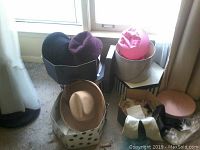 Photo of multiple ladies hats in various colors and styles placed on and around chairs and boxes.