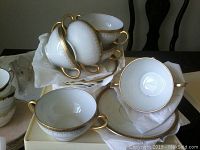 Stack of white Limoges sharing teacups with gold handles and gold trimmed rims on and near white saucers with matching gold decorative edge patterns.