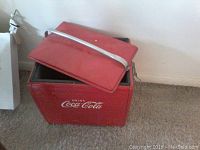 Red Coca-Cola cooler with open hinged lid showing interior lining and metal handle on the lid, placed on carpet.