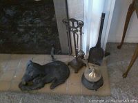 Close view of metal dog statue lying on tiled floor with fireplace tool stand and brass pot behind it.