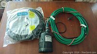 Mastercraft submersible water pump, coiled green electrical cord, and Burke flexible hose in packaging on wooden table.