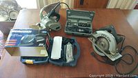 All four power tools arranged on a table including the belt sander, detail sander in box, rotary tool case, and SkilSaw circular saw
