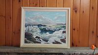 Frontal view of original oil painting showing ocean waves, rocky shore, and frame against wooden wall.