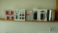 Photo of 6 boxes of Bodum tea and coffee items lined on a shelf, including glass mugs and French press boxes plus a tall metal container on the far right