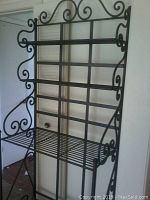 Full view of black iron shelving stand showing decorative scroll details and four wire shelves