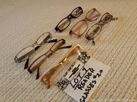 Seven pairs of assorted reader glasses with +2.0 lens strength, displayed on light fabric background.
