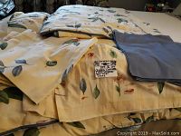 Stacked bedding and window valances with floral leaf print and solid blue-gray fabric, showing tags for queen comforter set.
