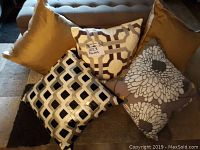 Set of five decorative pillows on carpet showing gold, cream with geometric pattern, cream and black lattice pattern, and gray floral pillow.