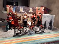 Overall display of six Christmas village figurines, iron fencing, small snowman, and boxed village accessories on a table with house backdrop.