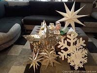Image showing summary of Christmas decorations on a gray ottoman against a dark sofa. Visible are large white snowflakes, a large paper star, a reflective glass star, smaller gold stars, and various small glass ornaments.