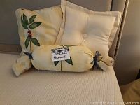 Three pillows arranged on a light beige upholstered seat: two square pillows - one with botanical print, one solid cream with tassel, and one bolster pillow with botanical print fabric tied at both ends with blue ribbons.