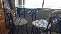 Round black glass pub table with two black iron framed chairs with white and black embroidered fabric upholstery on seat and back