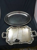 Photo showing two silver plated serving trays, one oval and one rectangular with handles, placed on a black background.