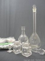 Lot of vintage glass items including a 250ml Pyrex volumetric flask, a clear etched glass vase, two small square glass inkwells with stoppers, and small glass animal figurines laid out on a gray surface.