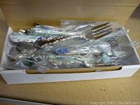 Box filled with multiple pieces of Community silver plated flatware wrapped in plastic bags, showing ornate handle designs and some tarnish.