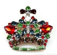 Close-up image of the colorful crown-shaped brooch with multicolored rhinestones mainly in green, red, blue, amber and clear on metal base.