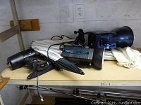 Photo showing various photography equipment including AMP FLASH 1000 units, umbrellas, and stands on a wooden shelf.
