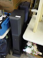 Pair of black JVC standing speakers with fabric-type grille covers front view, placed closely side-by-side near other stored items.