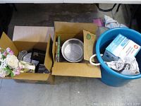 Two cardboard boxes filled with home and kitchen items, metal strainer visible in one box, a large blue tub with a box labeled Ultrasoft H inside and crumpled paper.