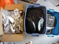 Three containers including a cardboard box full of assorted silver plated cutlery wrapped in plastic bags, a gray Rubbermaid tote holding several black speckled roasting pans with lids, and a blue Rubbermaid tote containing a George Foreman electric grill with power cord.
