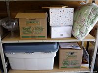 Multiple cardboard boxes and a large plastic tote, containing various housewares including linens, scarves, glassware, and ceramics packed inside.