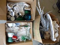 Photo of two cardboard boxes filled with assorted vintage houseware and decorative items including glass jars, ceramic cups, metal kitchen utensils, and a large decorative plate
