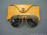 Vintage Bushnell aviator sunglasses with gold-toned metal frames and dark lenses lying in front of the brown leather case.