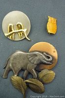 Group of 3 animal themed brooches and pins including elephant brooch, bird pin, and owl pin, on a dark textured background.