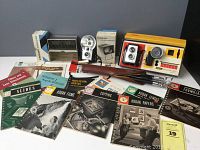 Photo showing the two Brownie cameras with one in original box, 12x magnifying slide viewer in box, tripod case, slide magazines, and 12 vintage photography books arranged on a table.
