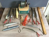 Full view of 13 saltwater fishing lures laid flat on table with ruler and Pflueger Capitol reel on top of its box showing scale and components.